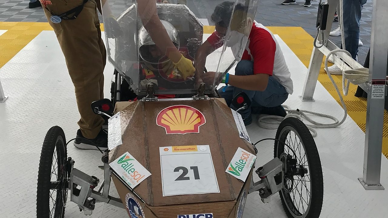 joven adulto en el interior de un coche de carreras, con el casco puesto, mientras es asistido por dos hombres para montar su equipo previo a la carrera. El coche de carreras es de color marrón, con el número 21 escrito y el logotipo de Shell resaltado.
