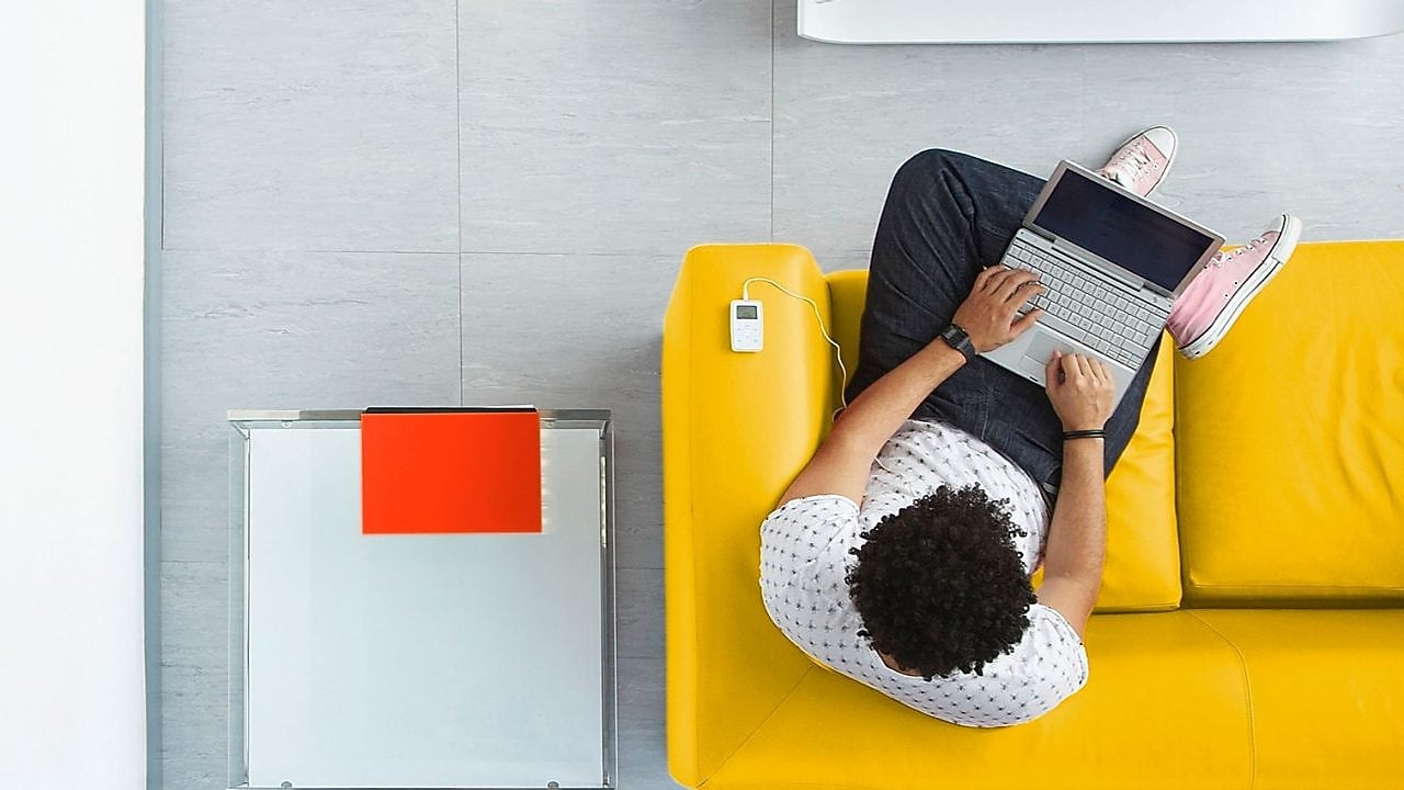 un hombre sentado en un sofá amarillo jugueteando con un laptop.