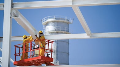 dos empleados con equipo de seguridad amarillo en una grúa roja, moviendo una viga blanca.