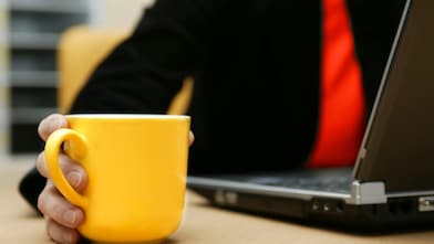 Una mano aparentemente masculina sostiene una taza amarilla. Al fondo vemos que la persona lleva una camisa negra y una corbata roja.