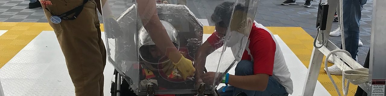joven adulto en el interior de un coche de carreras, con el casco puesto, mientras es asistido por dos hombres para montar su equipo previo a la carrera. El coche de carreras es de color marrón, con el número 21 escrito y el logotipo de Shell resaltado.