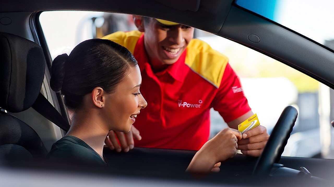 mujer al volante, en un coche parado, siendo atendida por un empleado de una gasolinera Shell. Tiene el pelo negro y sonríe, lleva uniforme y una gorra roja. Ella le enseña una tarjeta Shell.