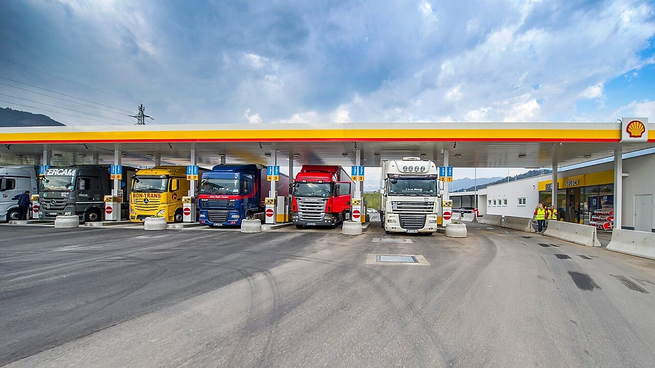 Gasolinera Shell con seis camiones aparcados. De izquierda a derecha: un camión blanco, verde oscuro, amarillo, azul oscuro, rojo y otro camión blanco.